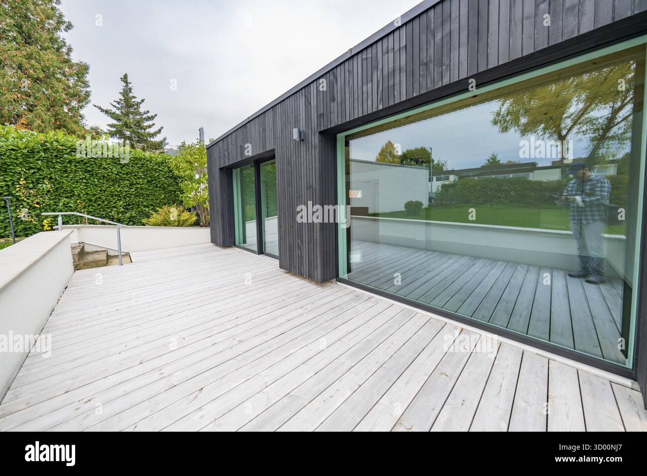 Minimalistischer Anbau mit schwarzer Holzfassade und großer Glasfront öffnet sich auf eine breite Holzterrasse im ruhigen Garten, Hausrenovierung, Klimaanlage Stockfoto