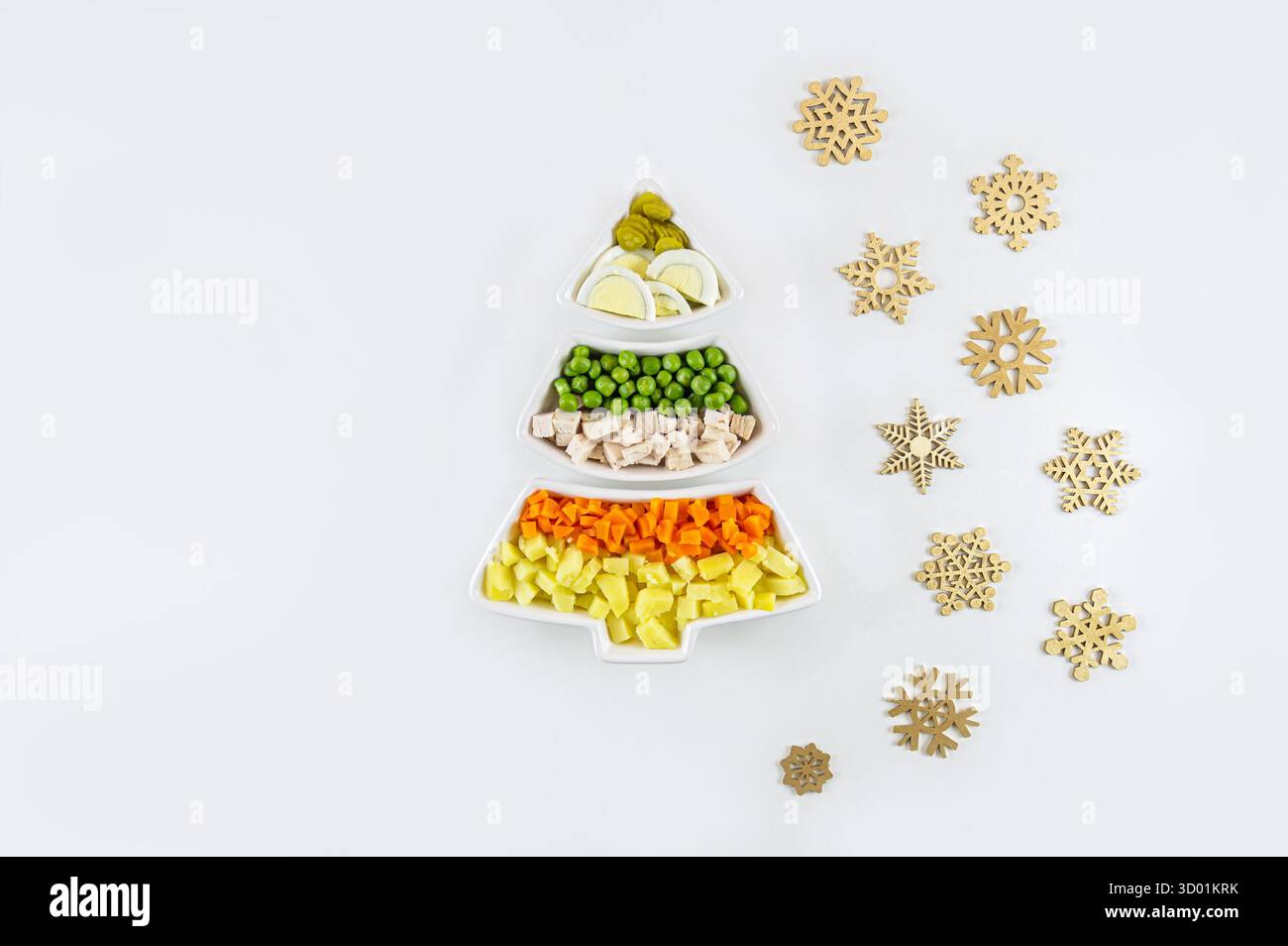 Die Zutaten für den olivier-Salat sind in weißen Schüsseln angeordnet, die wie ein Weihnachtsbaum auf weißem Hintergrund mit Holzschneeflocken geformt sind. Festlich, minimalistisch Stockfoto