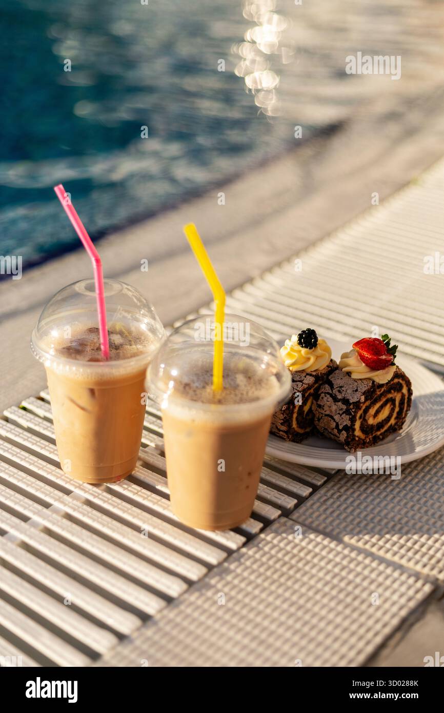Zwei Eiskaffee-Drinks mit bunten Strohhalmen sitzen an einem hellen, sonnigen Nachmittag neben einem Teller mit Schokoladenrollen in der Nähe eines glitzernden Pools. Stockfoto