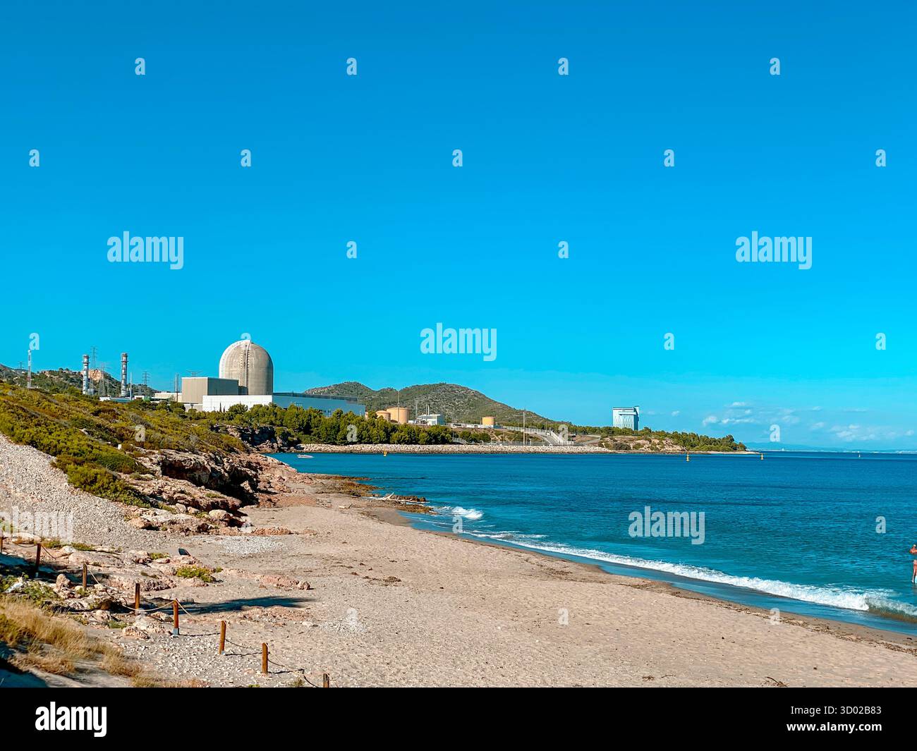 Strand La Almadrava in Hospitalet del Infant, Spanien, mit dem Kernkraftwerk Vandellos II im Hintergrund, ruhigem blauem Meer und sonnigem, klarem Wetter Stockfoto
