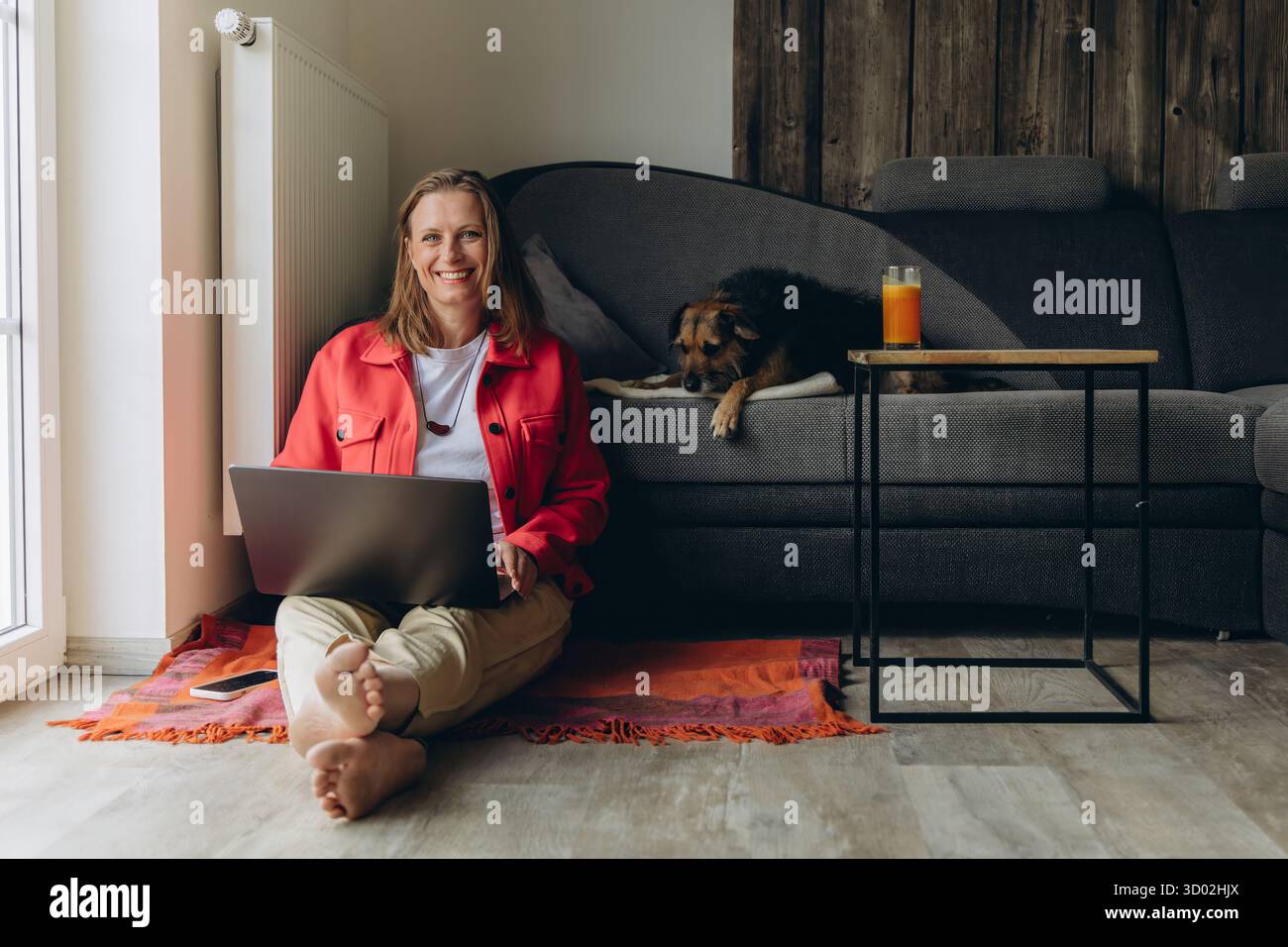 Frau, die zu Hause mit Laptop arbeitet, barfuß und entspannt auf dem Boden in der Nähe des Fensters sitzt, mit Hund an der Seite. Gemütlicher freiberuflicher Lebensstil und Stockfoto