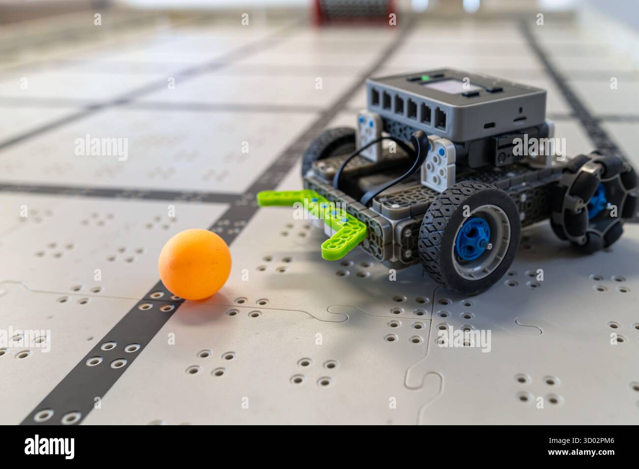 Ein kleines Roboterauto bewegt sich auf einem gemusterten Boden in einer technischen Umgebung auf eine leuchtende orangefarbene Kugel zu. Stockfoto