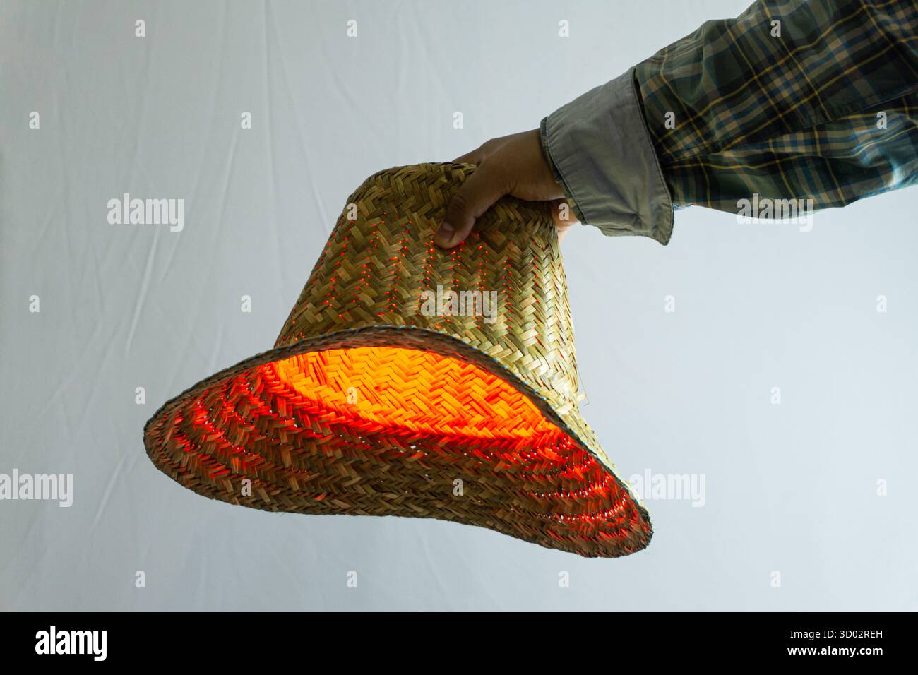 Strohhut in der Hand gehalten mit rotem Licht, das von der Oberfläche reflektiert wird, traditionelles Accessoire in dramatischem Studio, aufgenommen auf weißem Hintergrund mit dezenter Atmosphäre Stockfoto