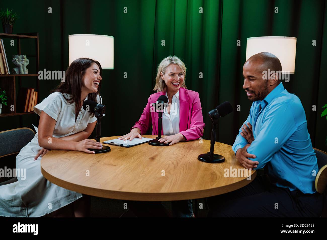 Drei verschiedene Erwachsene Gastgeber, ein Mann und zwei Frauen, die mit Mikrofonen an einem Tisch sitzen, an einer lebhaften Podcast-Diskussion teilnehmen oder ein Interview in einem Cur führen Stockfoto
