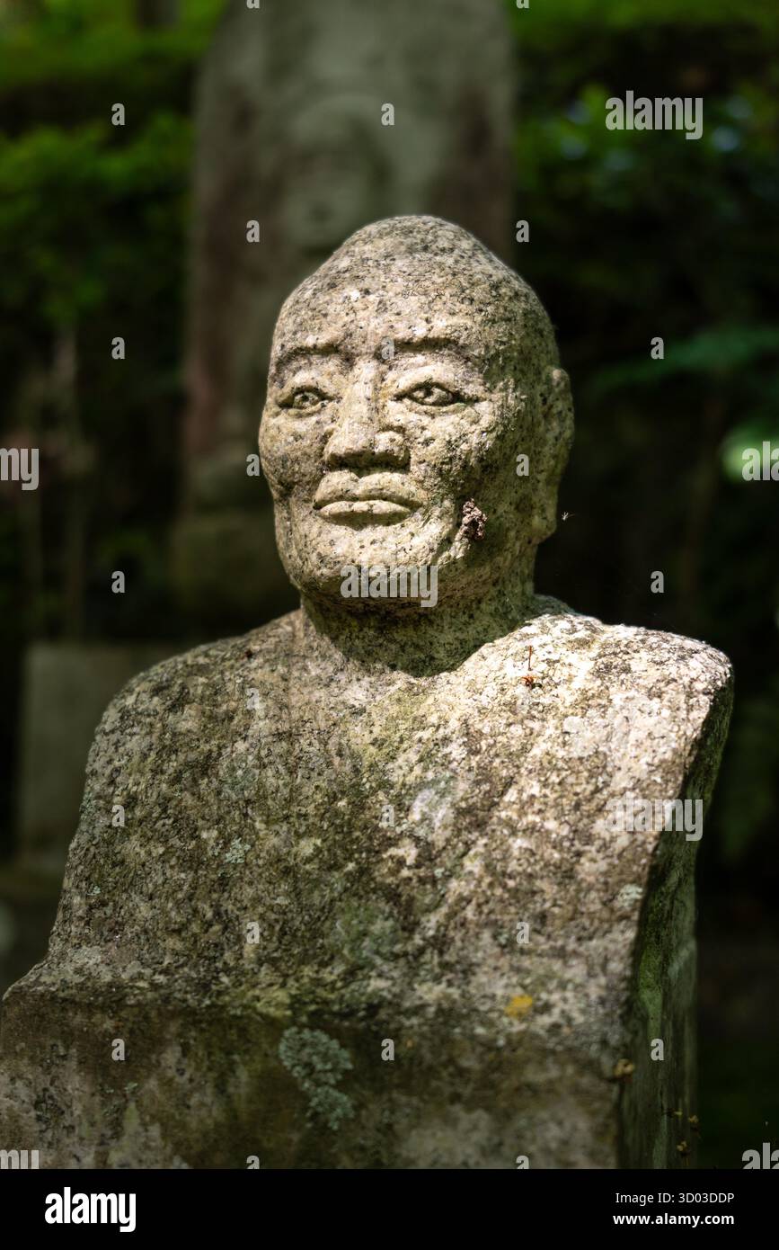 Eine verwitterte Steinstatue einer ruhigen Figur steht in ruhiger Betrachtung am Mitaki-Dera Tempel in Hiroshima, umgeben von Waldschatten Stockfoto Eine verwitterte Steinstatue einer ruhigen Figur steht in ruhiger Betrachtung am Mitaki-Dera Tempel in Hiroshima, umgeben von Waldschatten Stockfoto