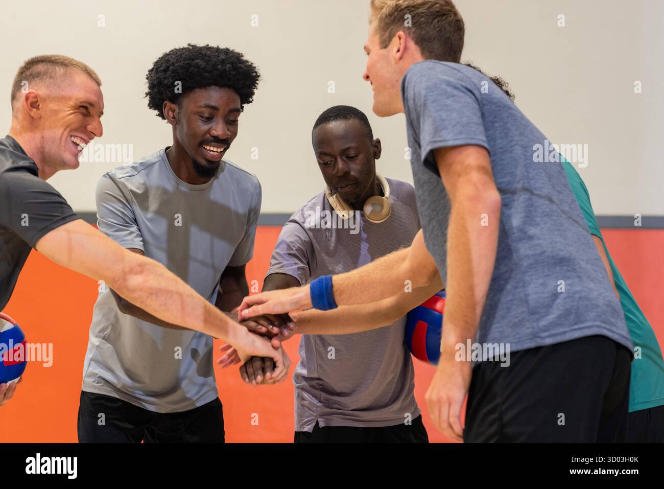 Verschiedene männliche Mannschaftskameraden stapeln Hände in der Sporthalle, halten Volleybälle und tragen Kopfhörer. Athleten, Teamwork, Training, Vibrant, Kameradschaft, Fitness, Wettbewerb || Model veröffentlicht Stockfoto
