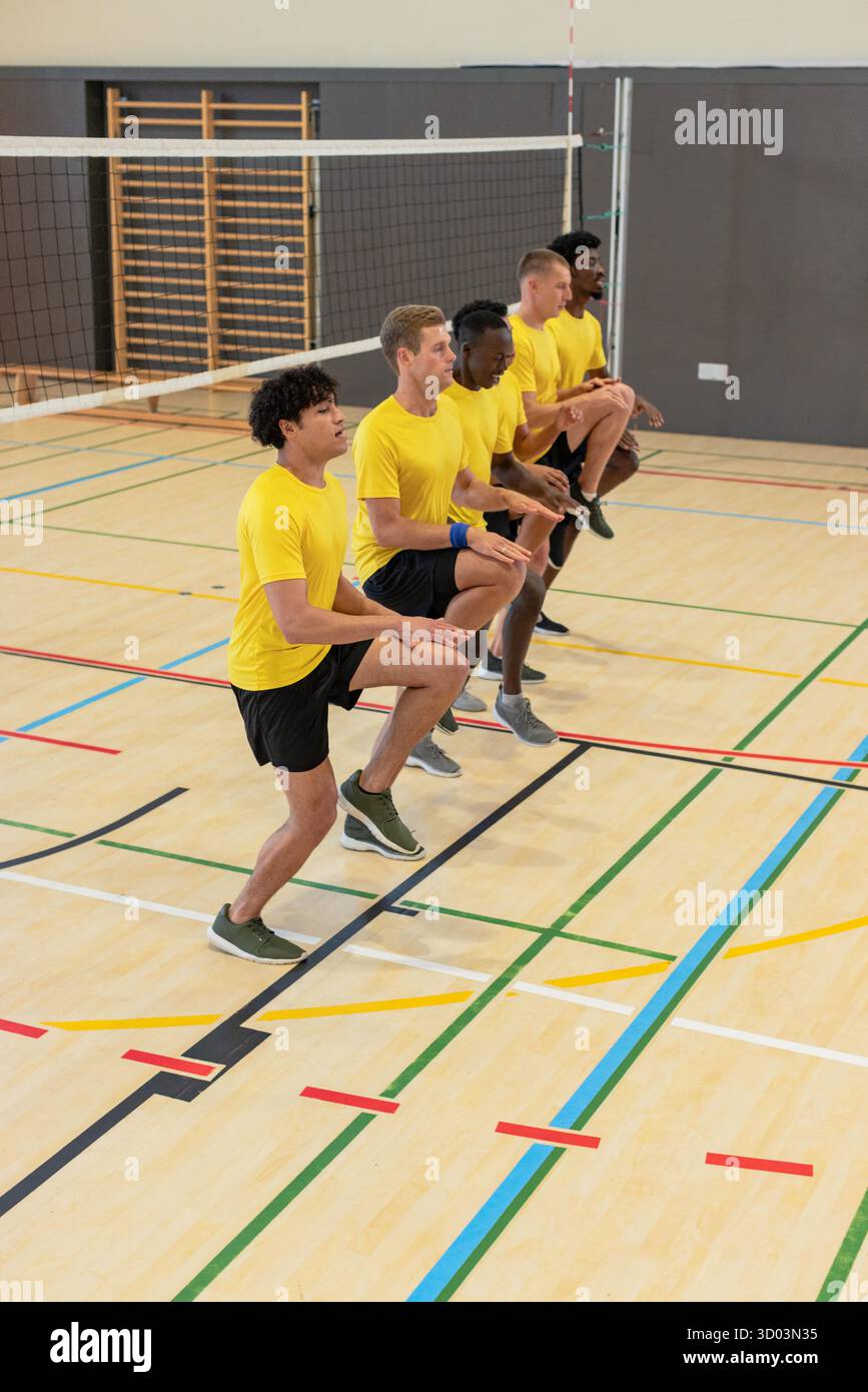 Verschiedene männliche Mannschaftskameraden führen synchronisierte Kniehegelungen in der Sporthalle mit Volleyballnetzpfosten durch. Athleten, Teamwork, Fitness, Training, Gymnasium, Übung, Kameradschaft || Model veröffentlicht Stockfoto