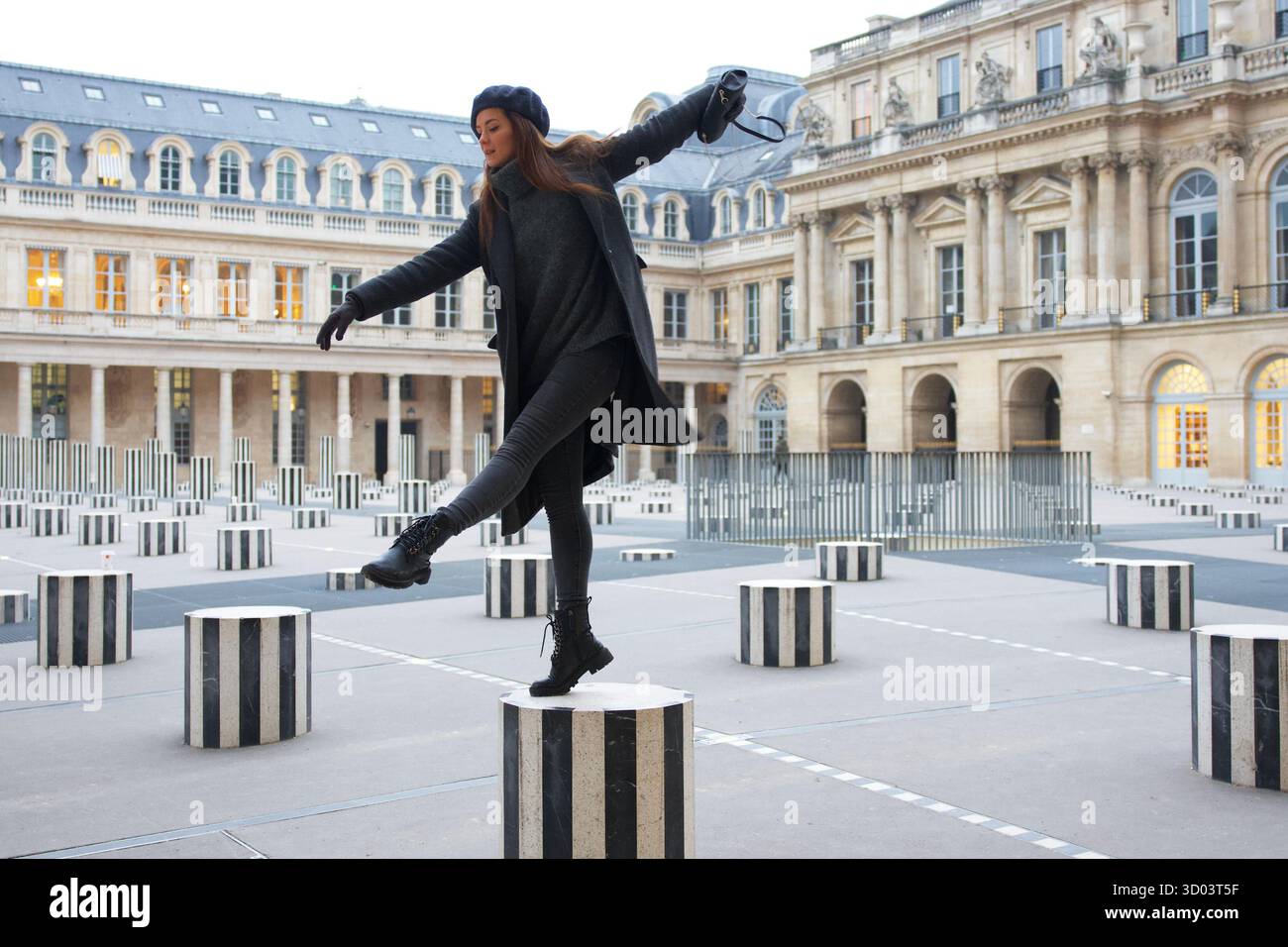 Eine Frau in Winterkleidung balanciert auf den gestreiften Säulen von Les Colonnes de Buren im Palais Royal. Ihre verspielte Pose verleiht dem eleganten Pariser Innenhof eine dynamische Note. Hochwertiges Foto || Modell veröffentlicht Stockfoto