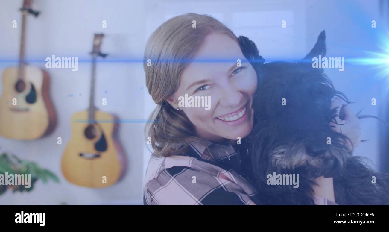 Lächelnde Erwachsene Frau, die einen kleinen schwarzen Hund im Wohnzimmer umarmt, mit Akustikgitarren und Pflanzen Stockfoto