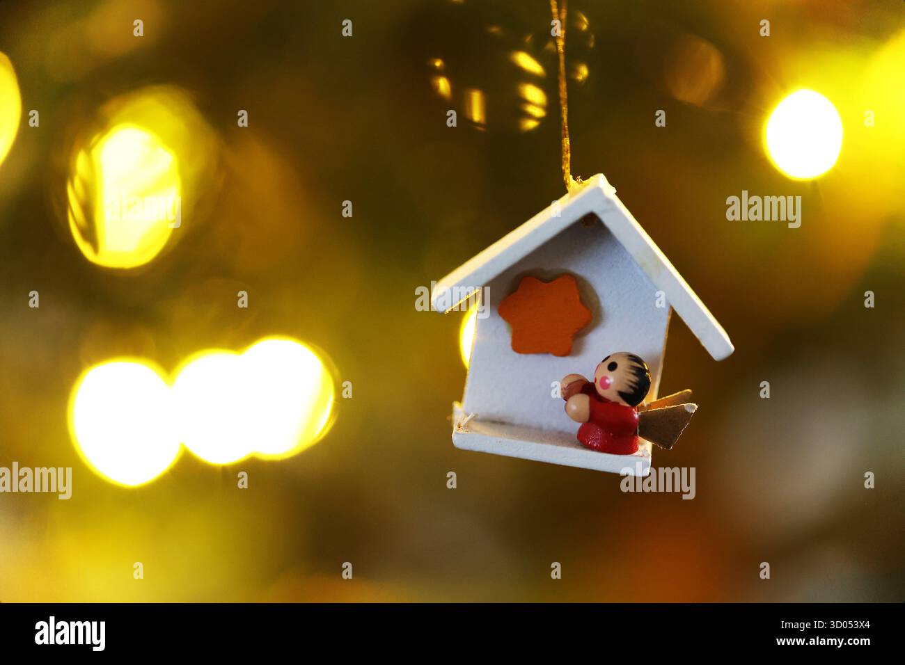 Weißes Miniatur-Haus-Ornament aus Holz mit einer Person im Inneren, die an einem Weihnachtsbaum mit warmen Bokeh-Lichtern im Hintergrund hängt Stockfoto