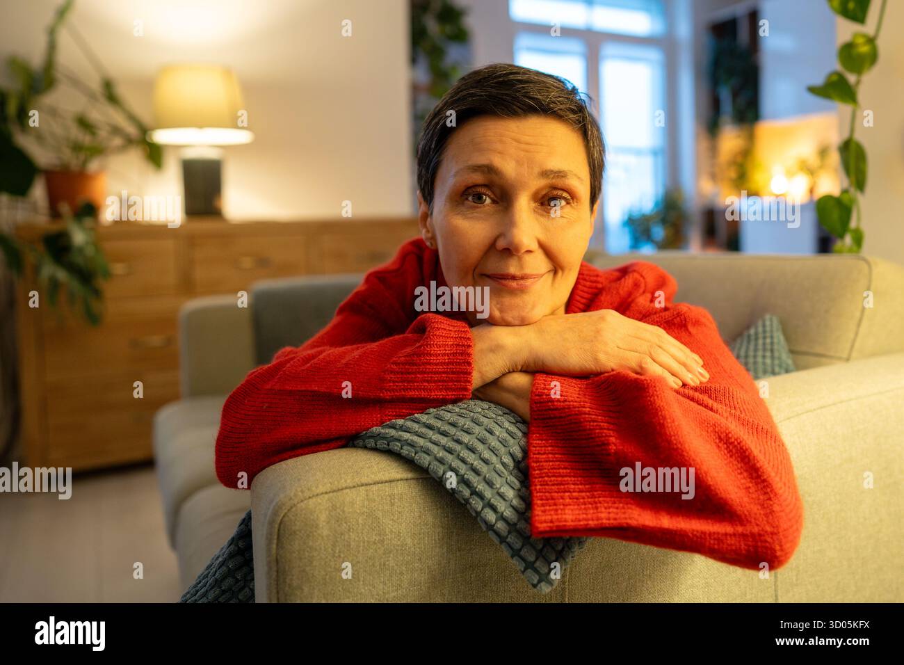 Lächelnde reife Frau, die Kopf auf Händen ruht. Glückliche, weibliche Kamera auf dem Sofa, anmutiger Ruhestand Stockfoto