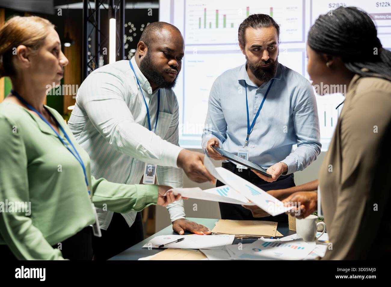 Multiethnische Teams tauschen Dokumente aus und nutzen die Datenvisualisierung, um Geschäftsziele in einem ruhigen Strategieraum zu prognostizieren. Mitarbeiter arbeiten spät in der Nacht zusammen. Stockfoto