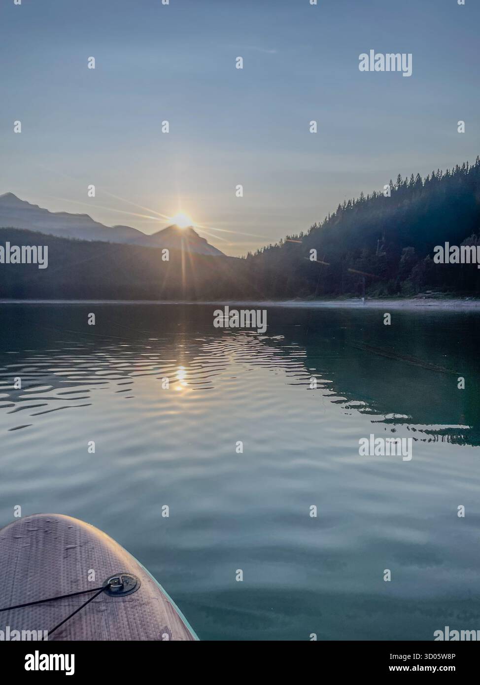 Eine ruhige Morgenszene auf einem klaren See, während ein Paddleboard in der Nähe des Ufers gleitet. Die Sonne geht hinter bewaldeten Hügeln auf und reflektiert goldenes Licht auf dem See - Smartphone-aufgenommenes Stockfoto