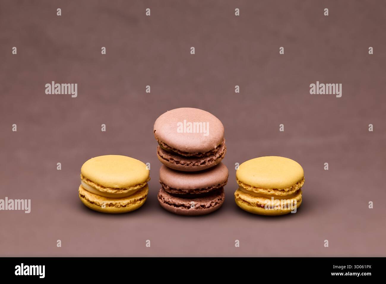Drei Macarons in Schokoladen- und Gelbtönen auf braunem Hintergrund in Studiobeleuchtung. Die minimale Einrichtung hebt Textur und Farbkontrast hervor Stockfoto