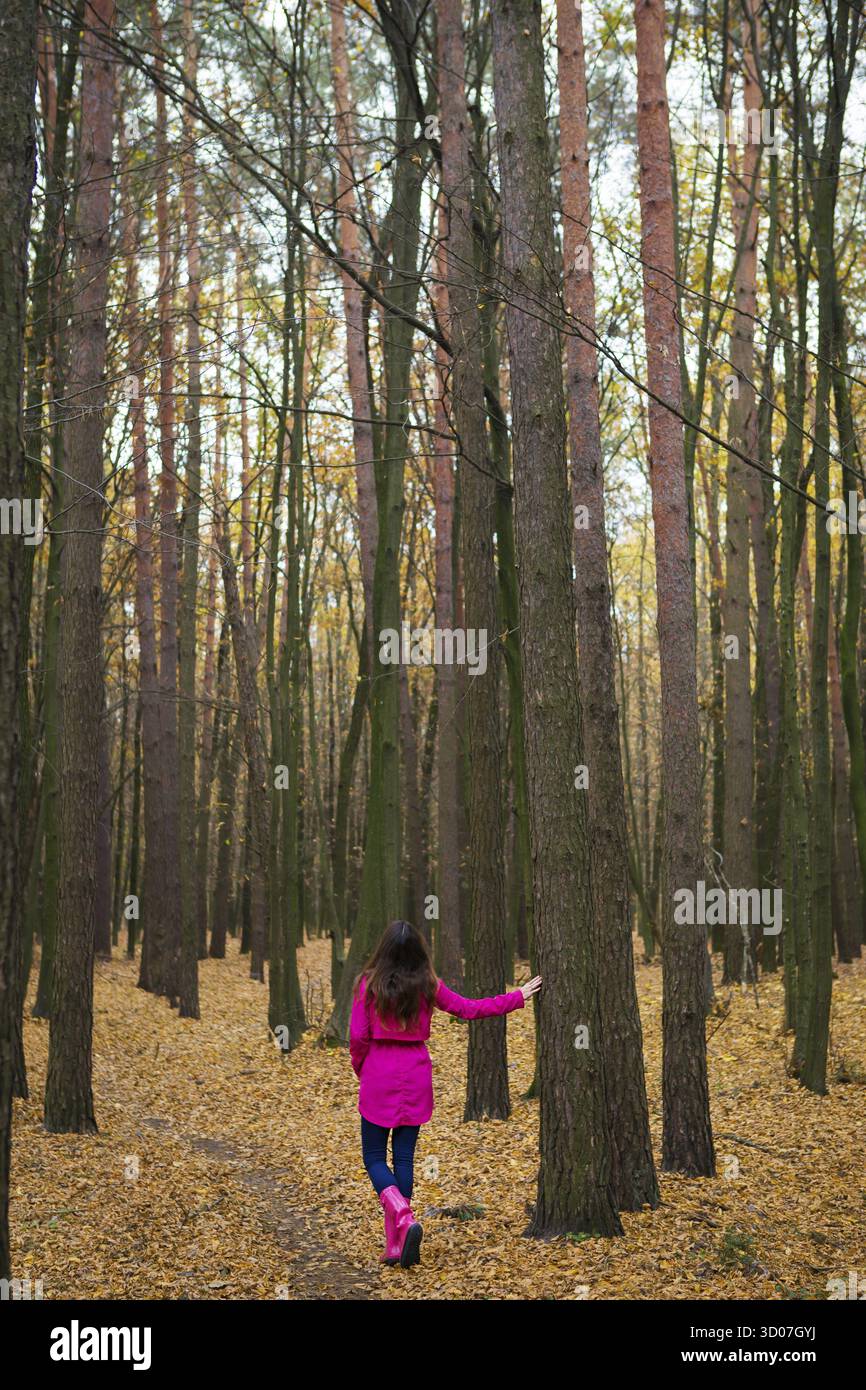 Mädchen in Regenmantel und rosa Gummistiefeln, die in einem Herbstwald spazieren Stockfoto