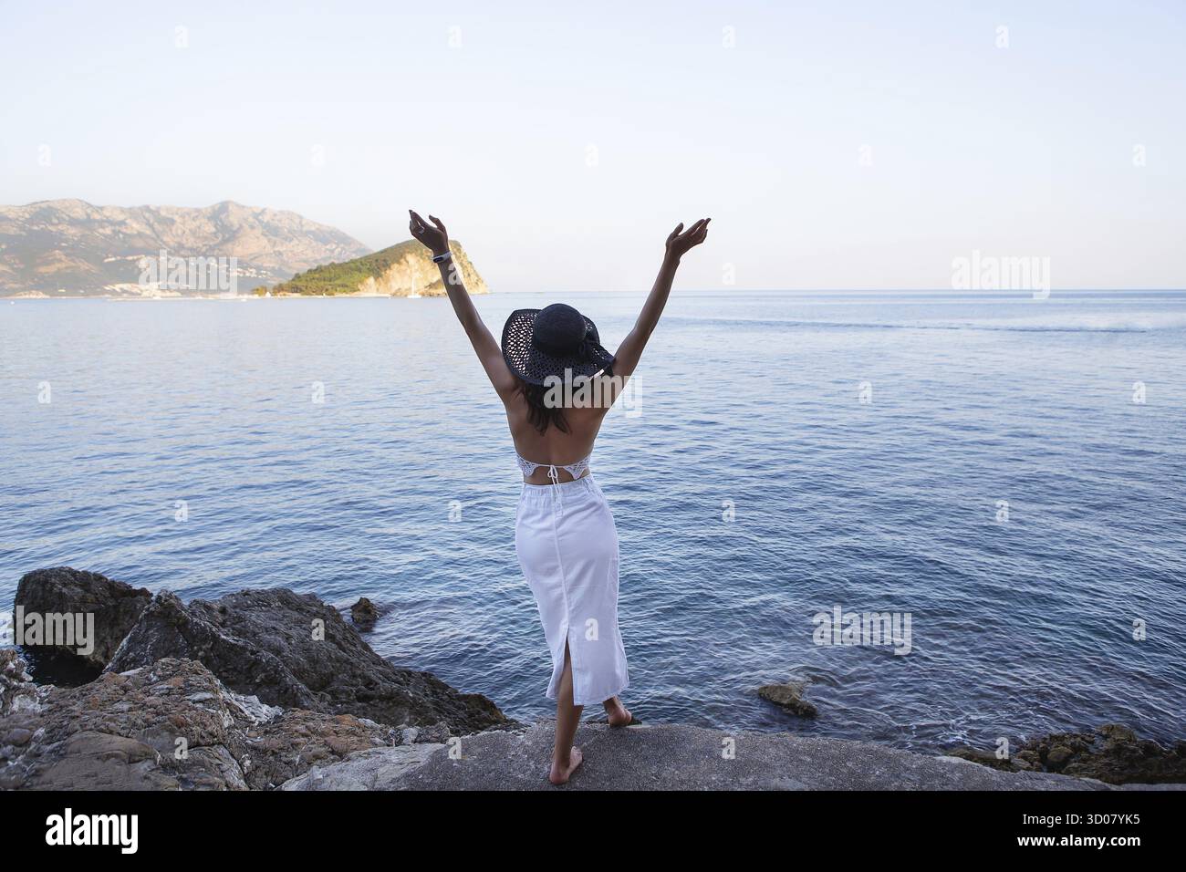 Eine Frau in weißem Kleid und Sonnenhut hebt freudig die Arme und blickt auf eine ruhige Meer- und Berglandschaft Stockfoto