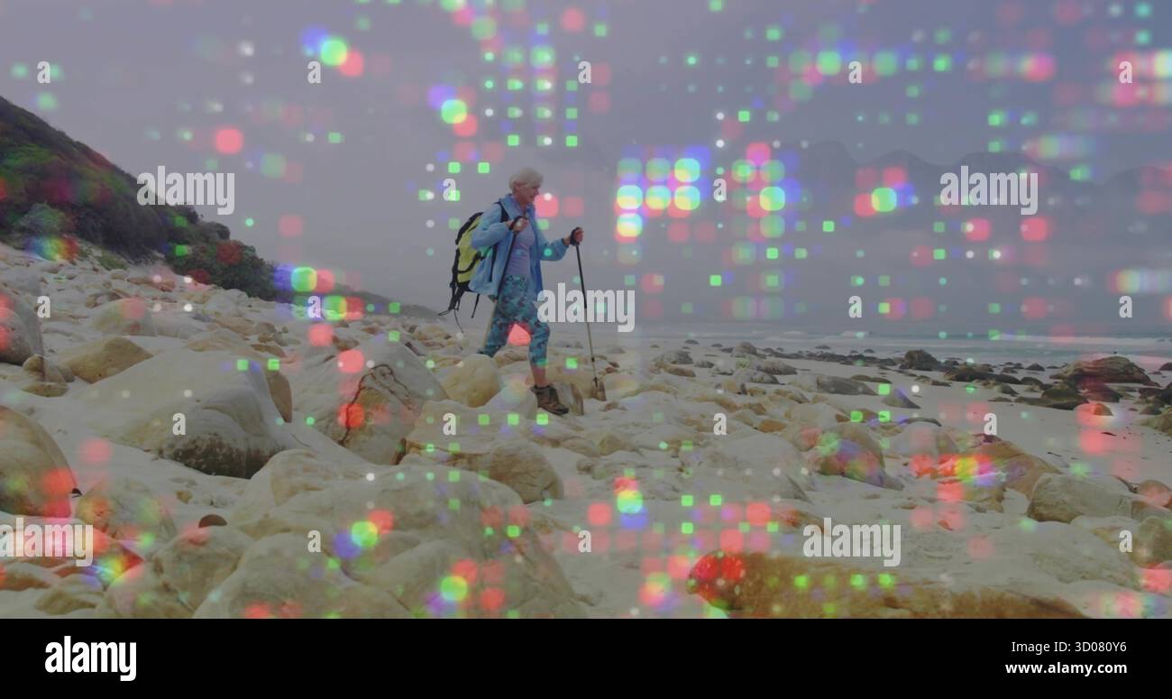 Eine ältere Frau in blauer Jacke, die die felsige Küste neben dem Meer navigiert, Wanderstöcke und Rucksack Stockfoto