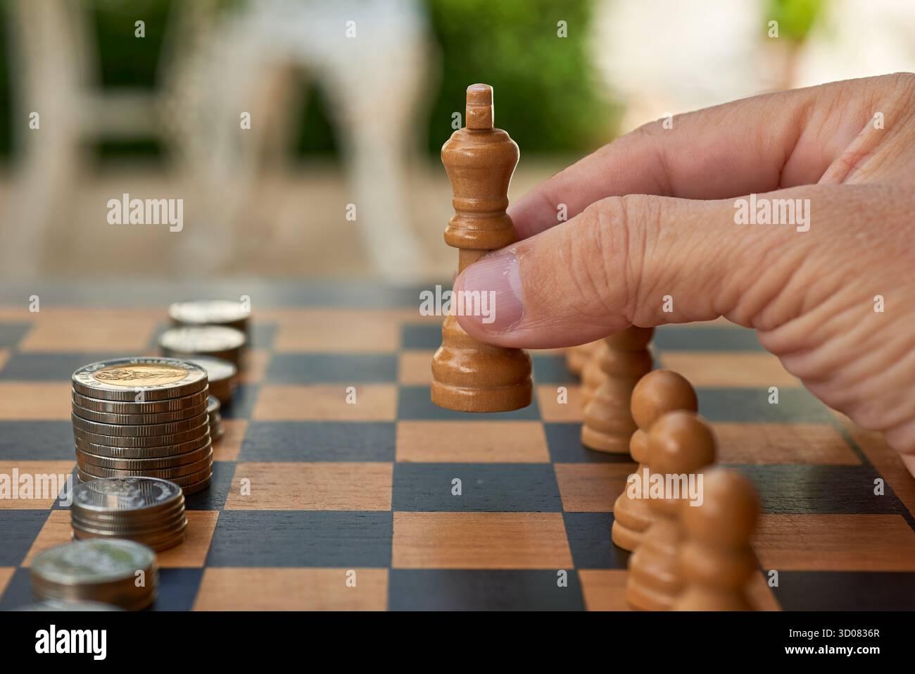 Eine Person greift nach draußen, um ein hölzernes Königsschach auf einem karierten Brett zu bewegen. In der Nähe befinden sich Stapel von Münzen, die eine entspannende und verächtliche Atmosphäre schaffen Stockfoto