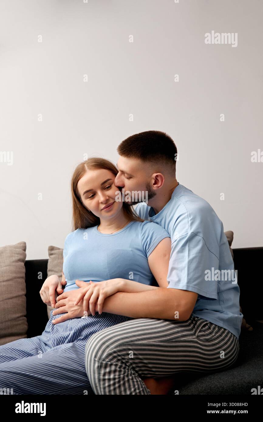 Das erwartende Paar genießt einen zärtlichen Moment auf der Couch, um ihre Liebe und die Erwartung zu feiern, ein neues Leben in ihrer Familie willkommen zu heißen Stockfoto