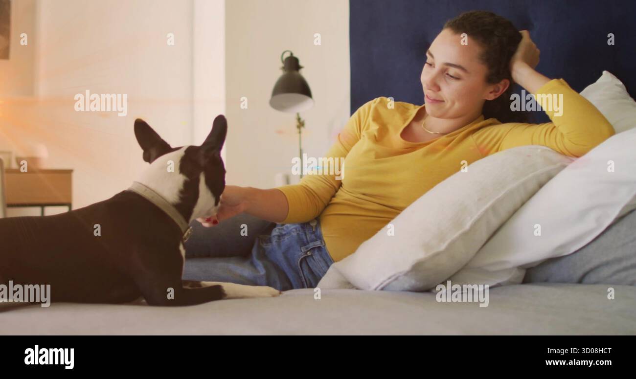 Liegende Frau mit Senfoberteil, die einen kleinen schwarzen und weißen Hund im Schlafzimmer streichelt, mit Leselampe Stockfoto