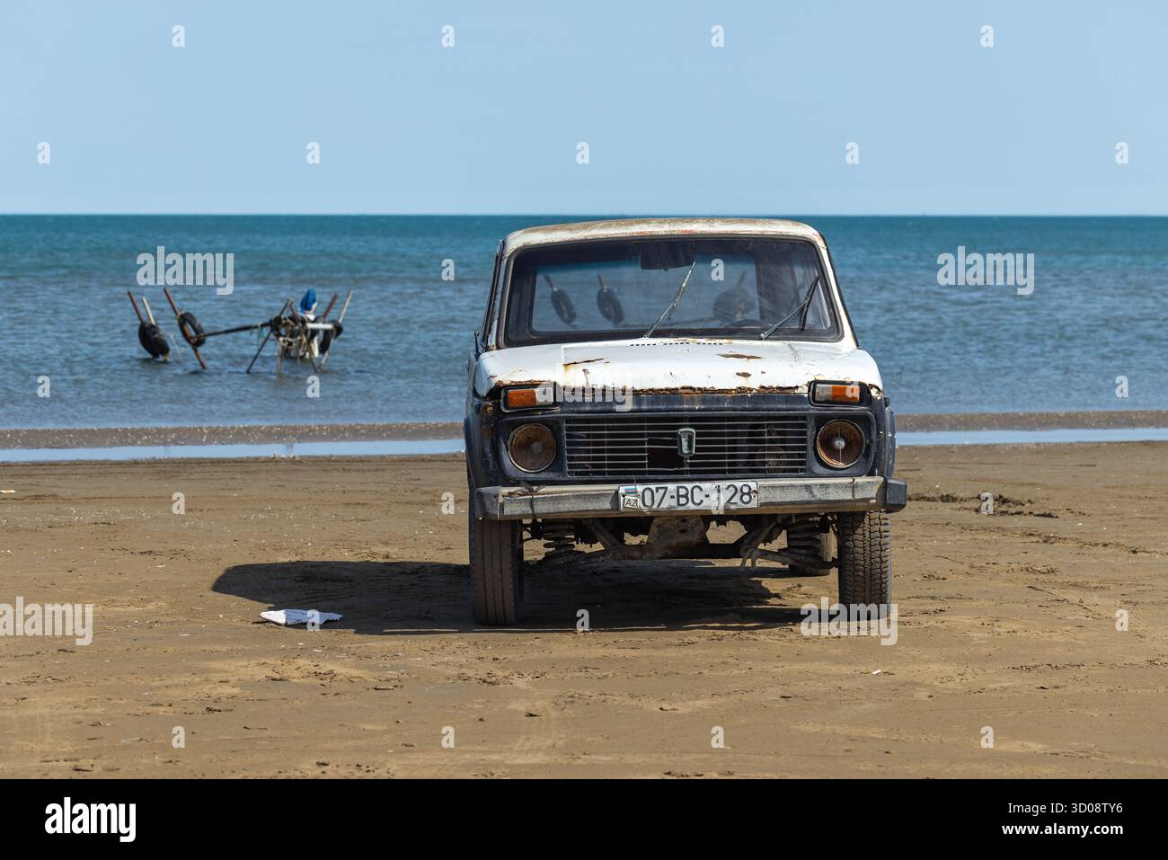 Buzowna, Aserbaidschan - 29. März 2025: Ein verwittertes weißes Vintage-Auto aus dem Jahr 1987 Lada Niva sitzt am Sandstrand, während blaue Wellen auf den Horizont treffen Stockfoto