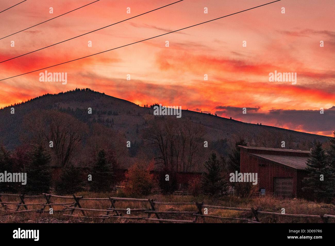 Stromleitungen einen rustikalen Holzzaun und ein älteres Nebengebäude der Farm unter einem dramatischen Sonnenaufgang über der Teton Range in den Rocky Mountains Stockfoto