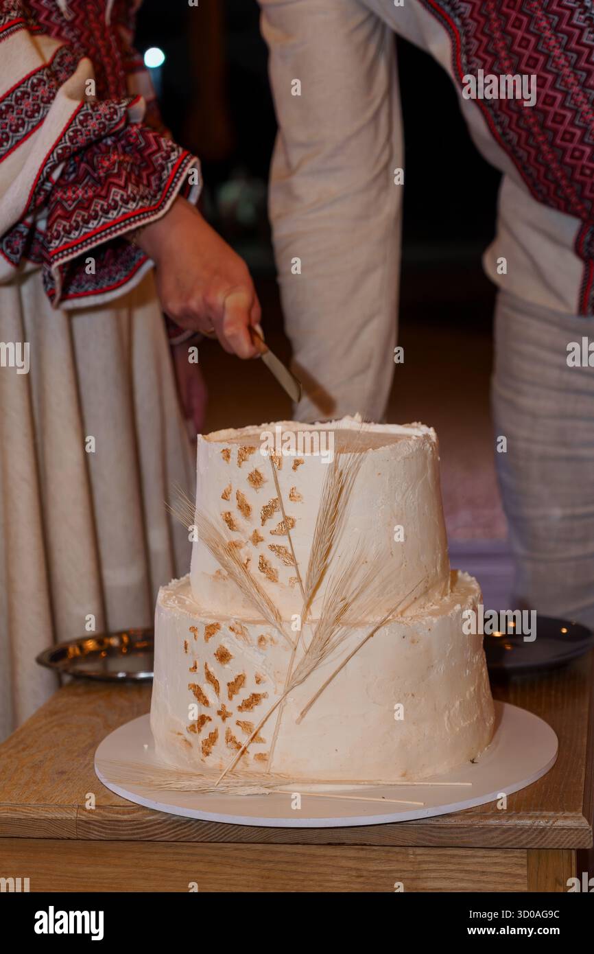 Eine Nahaufnahme einer traditionellen rumänischen Hochzeitstorte, die während einer Zeremonie geschnitten wird, mit komplizierten Volksstickereien auf der Kleidung der Partien Stockfoto