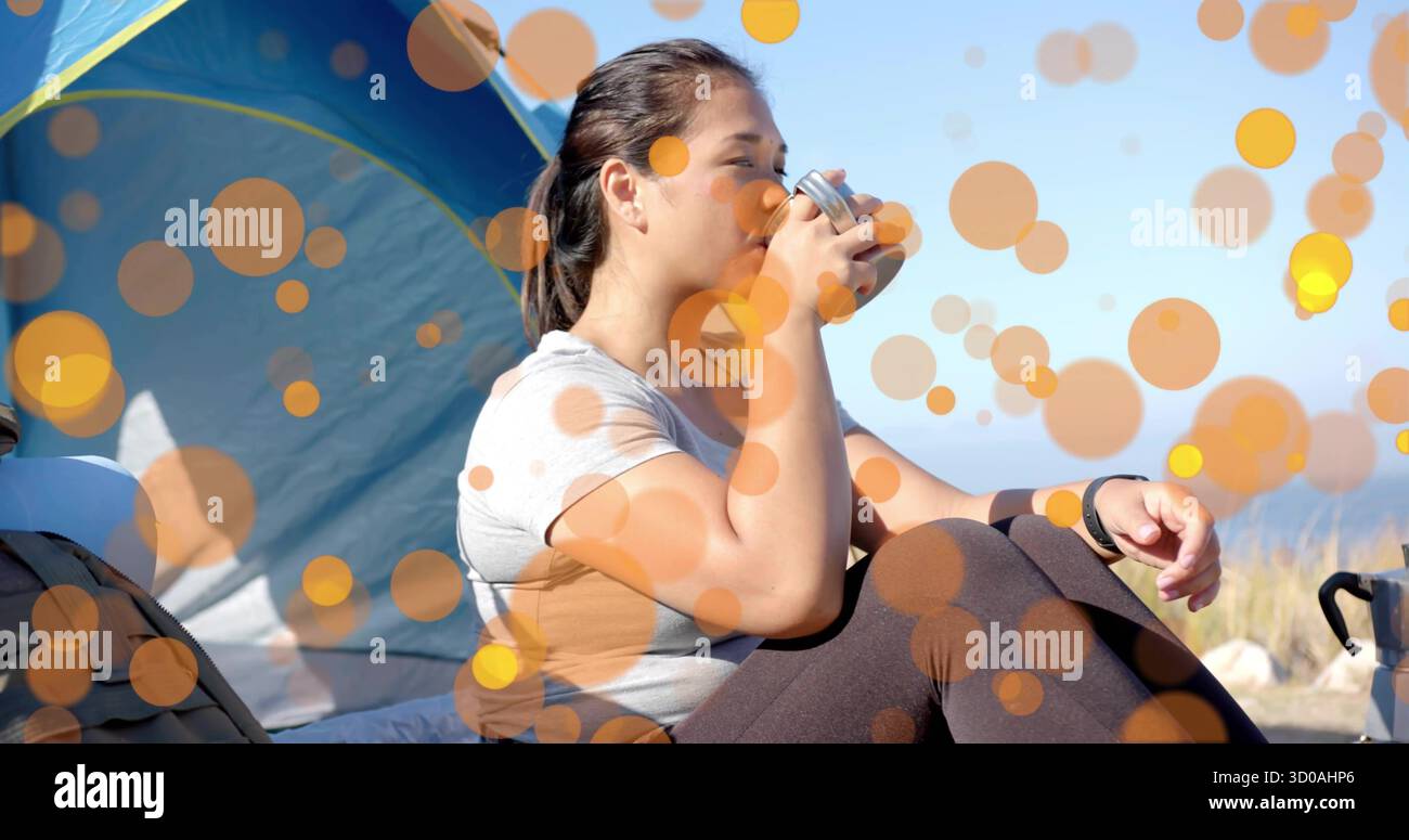 Eine trinkende Frau in Aktivkleidung sitzt auf dem Strandcamping und schlürft aus einem Metallbecher, mit blauem Zelt Stockfoto