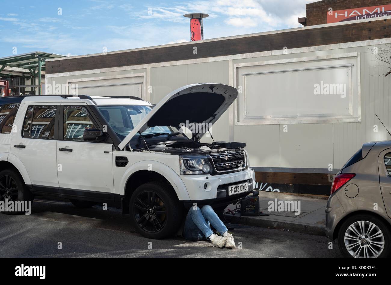 Ein Paar Beine eines Mechanikers, der unter dem Motor eines Land Rover arbeitet, der auf einer Wohnstraße in Hackney Wick, East London, Großbritannien, geparkt ist Stockfoto