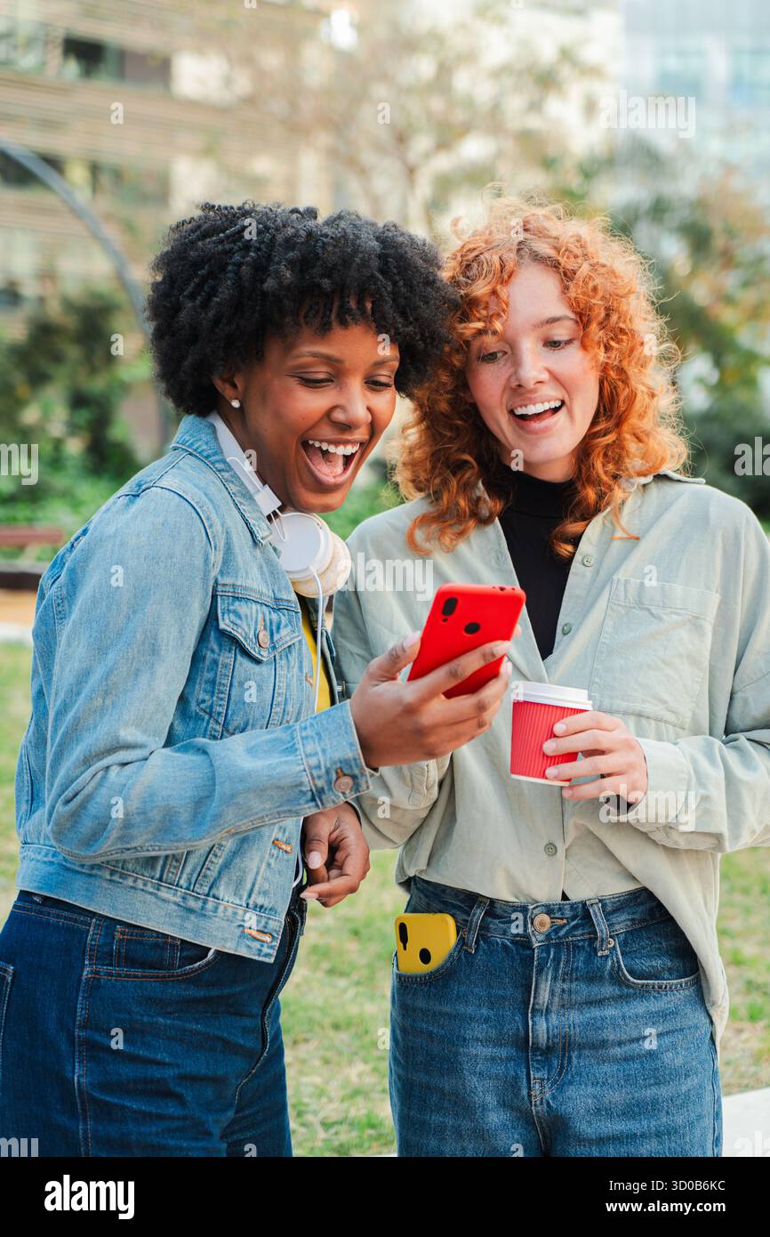Zwei begeisterte Frauen freuen sich über ein gemeinsames Erlebnis, während sie lachen und mit einem Smartphone interagieren und ihre Verbindung und Begeisterung zeigen Stockfoto