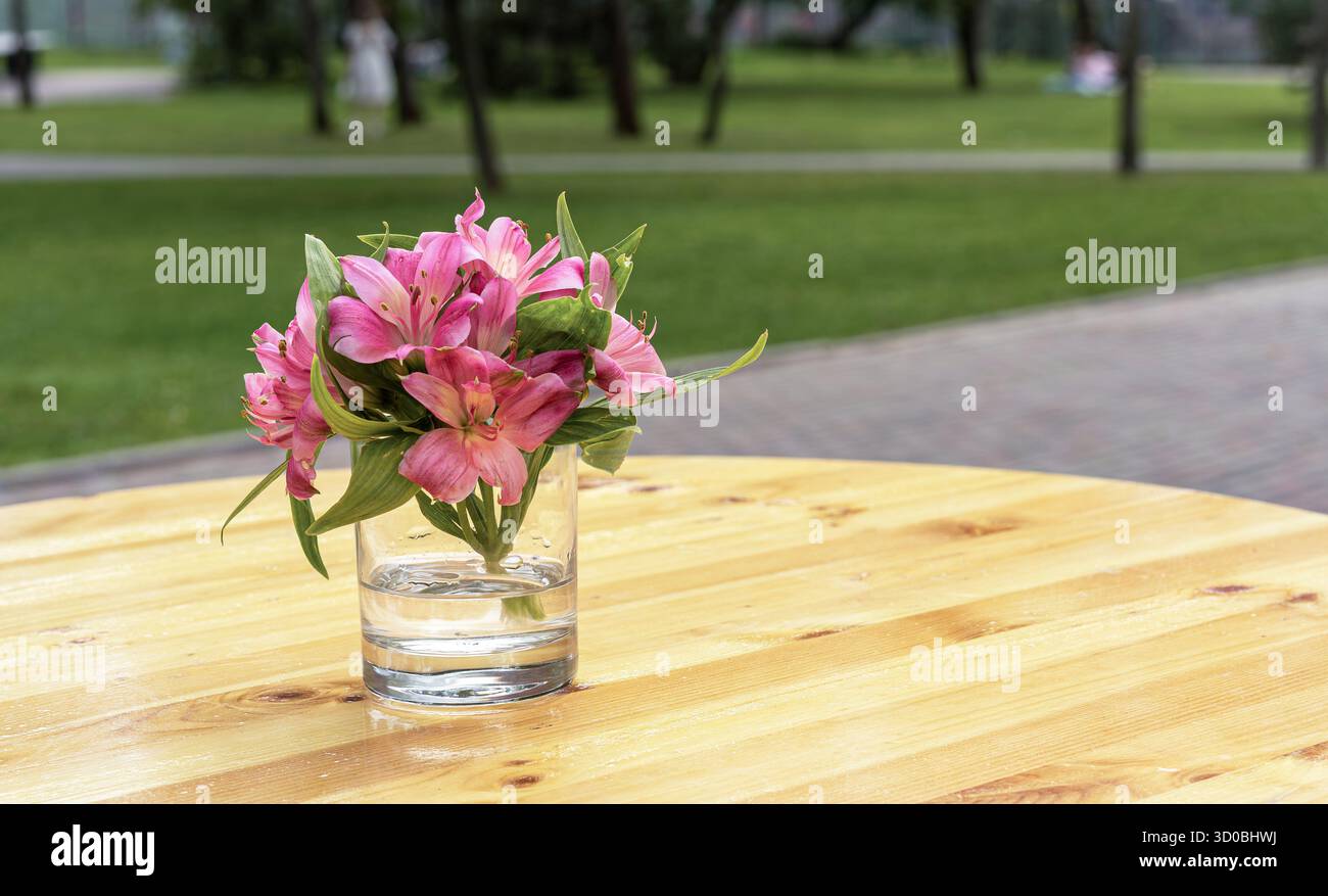 Zartes Bouquet rosa Lilien in Glasvase auf Holztisch mit verschwommenem Parkhintergrund, romantisches Stillleben im Freien Stockfoto