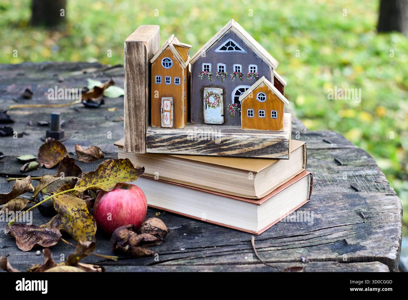 Stillleben im Herbst: Miniatur-Holzhäuser, Bücher, Apfel und gefallene Blätter auf einem alten Holztisch im Garten, die eine gemütliche saisonale Szene schaffen Stockfoto