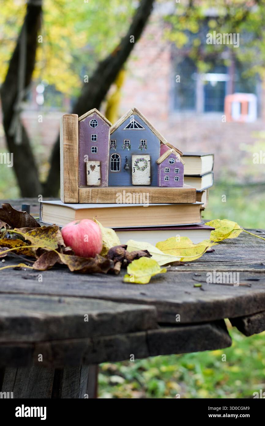 Gemütliches Stillleben im Herbst mit Büchern, einem Apfel und dekorativen Holzhäusern auf einem rustikalen Holztisch in einem Garten, die ein warmes Ambiente schaffen Stockfoto
