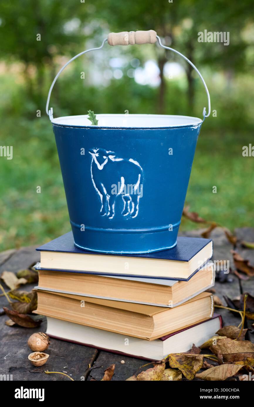 Blauer Emailleeimer mit einer bemalten Kuh auf einem Stapel Bücher und herabfallenden Herbstblättern, die eine gemütliche und rustikale Szene schaffen Stockfoto