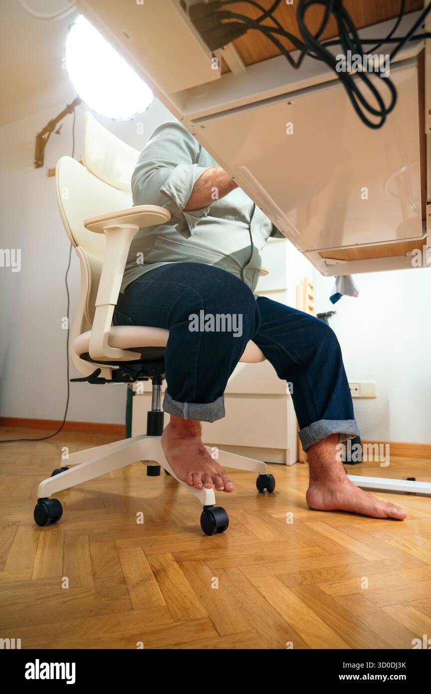 Barfuß sitzender Mann unter dem Schreibtisch auf ergonomischem Stuhl. Stockfoto