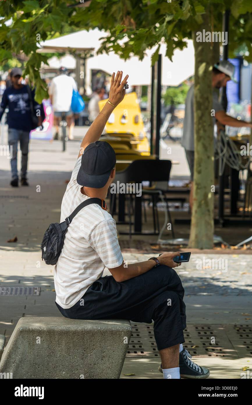 Ljubljana, Slowenien August 10-2025 Ein Mann sitzt auf einer Bank, der eine Hand hebt, um einen Freund zu begrüßen Stockfoto