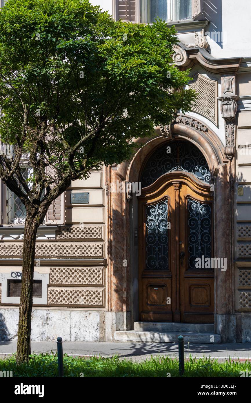Ljubljana, Slowenien 10. August 2025 kunstvoller Eingang geschnitzte Holztür und Steinfassade mit Baumschatten Stockfoto