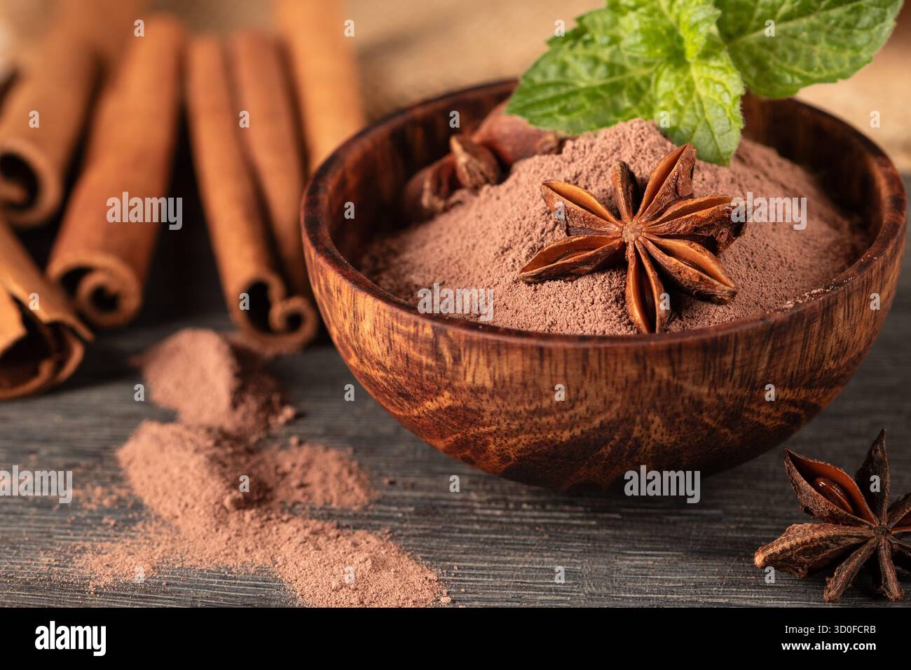 Diese Nahaufnahme zeigt eine rustikale Holztasse gefüllt mit reichhaltigem Kakaopulver, begleitet von duftendem Sternanis und warmen Zimtstäbchen, die kreieren Stockfoto