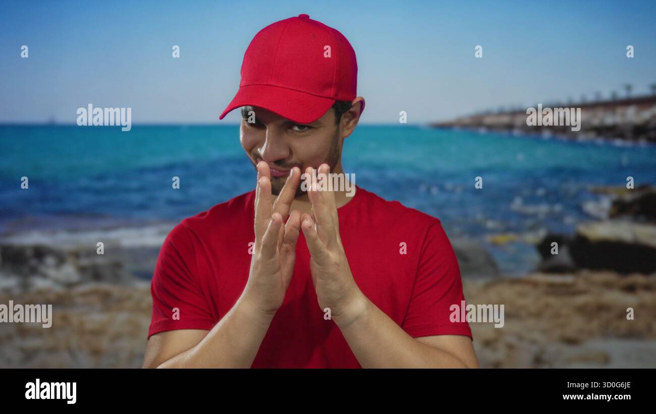 Junger hispanischer Kuriermann in roter Mütze und Hemd, mit den Fingern geschwenkt und mit einem leisen Grinsen an einem Strandpier. Stockfoto