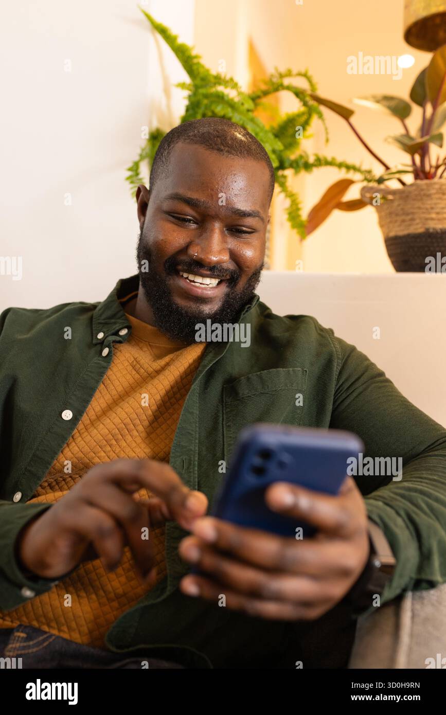 Afroamerikanischer Mann sitzt auf Sofa im Wohnzimmer und blättert Smartphone unter Umgebungslicht Stockfoto