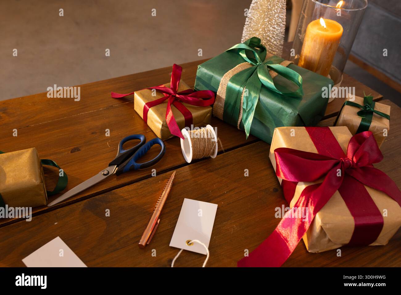 Schere und Garn liegen neben gewickelten grünen und goldenen Geschenken mit beleuchteter Kerze auf der Tischplatte Stockfoto