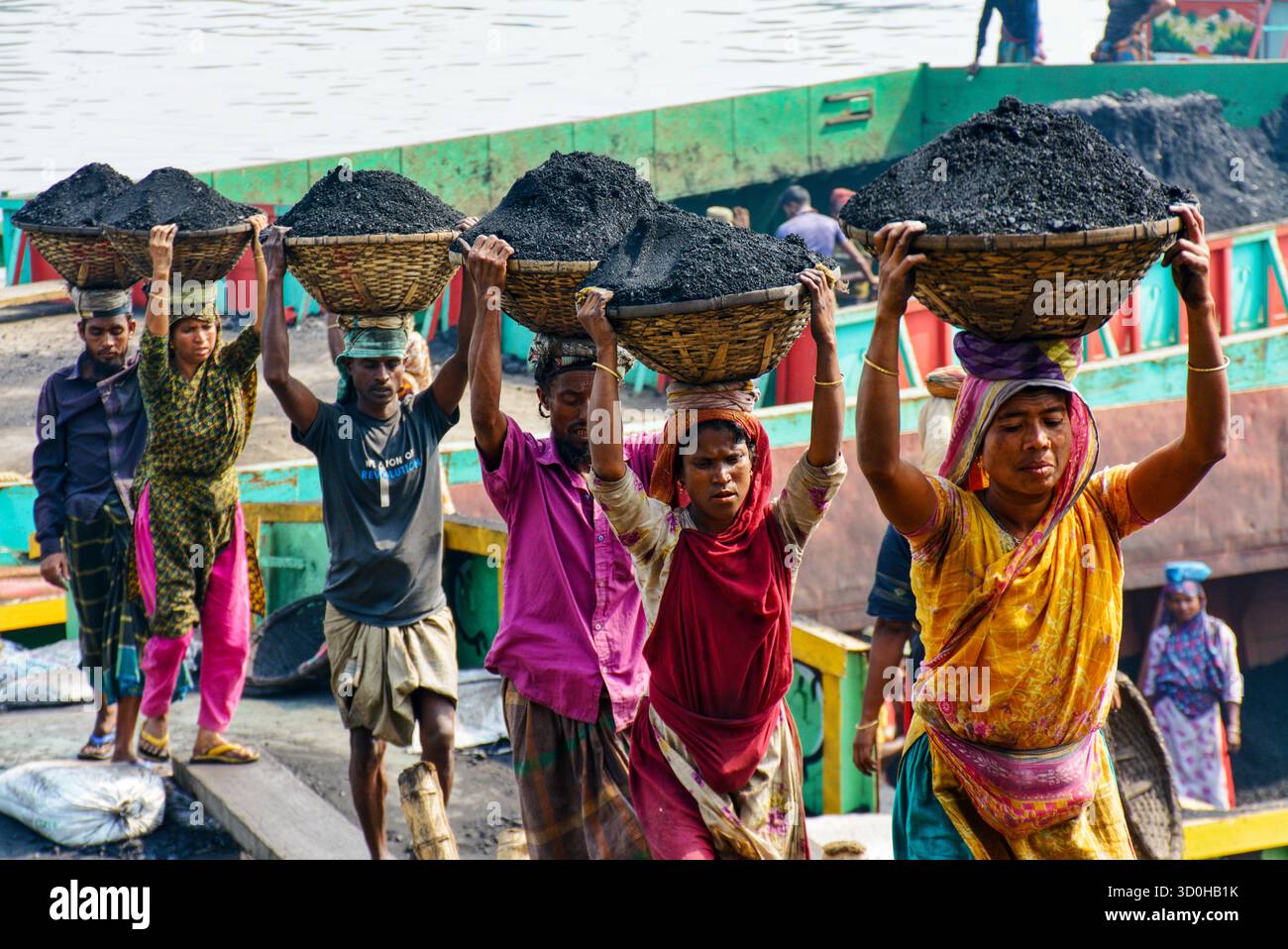 Dhaka, Bangladesch - 15. April 2024: Ein Blick auf Arbeiter, die schwere Mengen Kohle in gewebten Körben transportieren, eine düstere Szene, die die harte Realität der harten Arbeit vor dem Hintergrund industrieller Aktivitäten widerspiegelt. Stockfoto