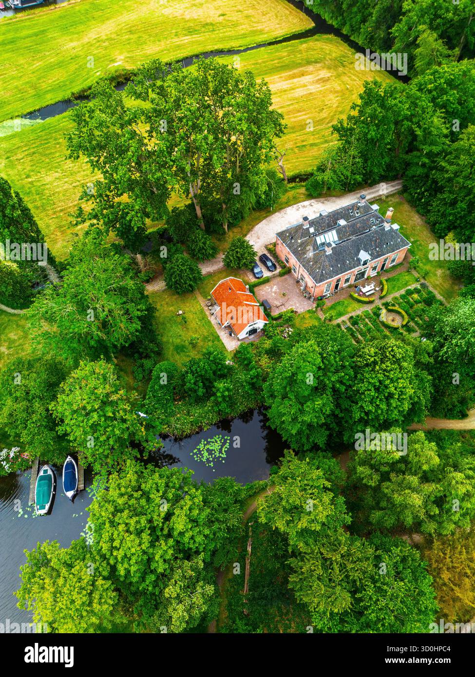 Blick aus der Vogelperspektive auf das Landgut Backsteinhaus mit dunklem Dach und rotem überdachtem Nebengebäude, umgeben von üppigem Grün, Kanal mit angedockten Booten und Gärten Stockfoto