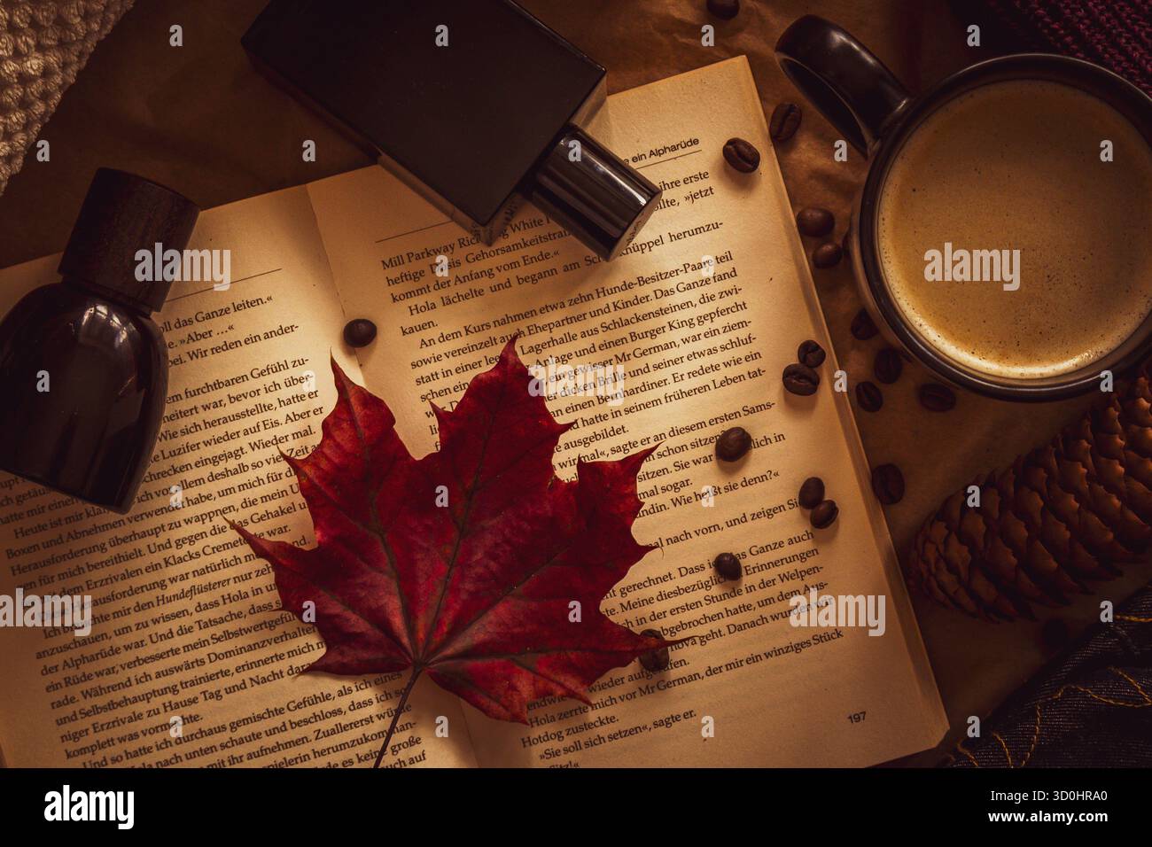 Gemütliches Herbstflachlegen mit offenem Buch, rotem Ahornblatt, einer Tasse Kaffee, Kaffeebohnen und Parfümflaschen auf warmem Hintergrund Stockfoto