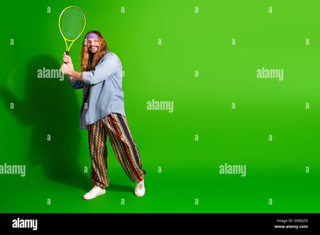 Junger Mann im Retro-Stil, der einen gelben Tennisschläger vor grünem Hintergrund hält und eine fröhliche und entspannte Haltung zeigt Stockfoto