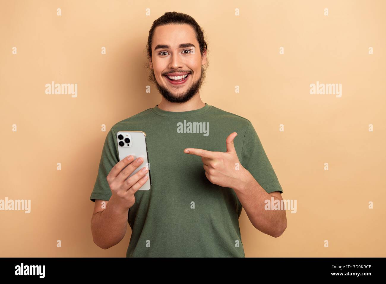 Glücklicher junger Mann mit Smartphone, das auf den Bildschirm zeigt und ein grünes T-Shirt vor beigefarbenem Hintergrund für Freizeit-Lifestyle-Shopping und Werbung trägt Stockfoto