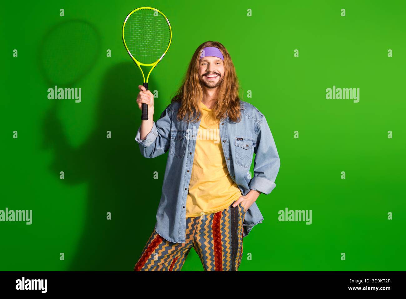Junger Mann mit langen Haaren im farbenfrohen Retro-Outfit, der Tennisschläger hält und vor einem leuchtend grünen Hintergrund lächelt Stockfoto