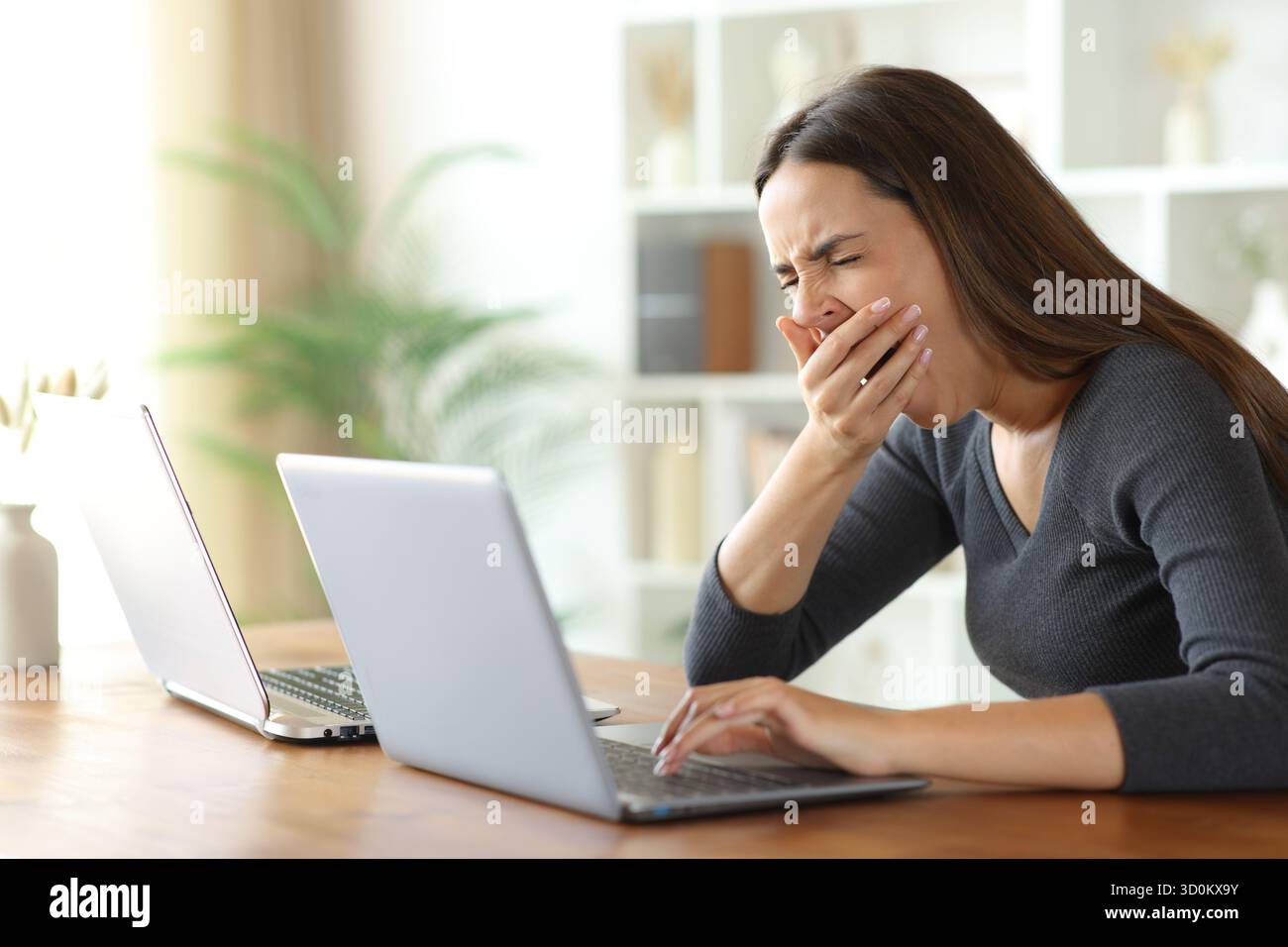 Müde Frau, die zwei Laptops auf einem Holztisch in einem Haus benutzt Stockfoto