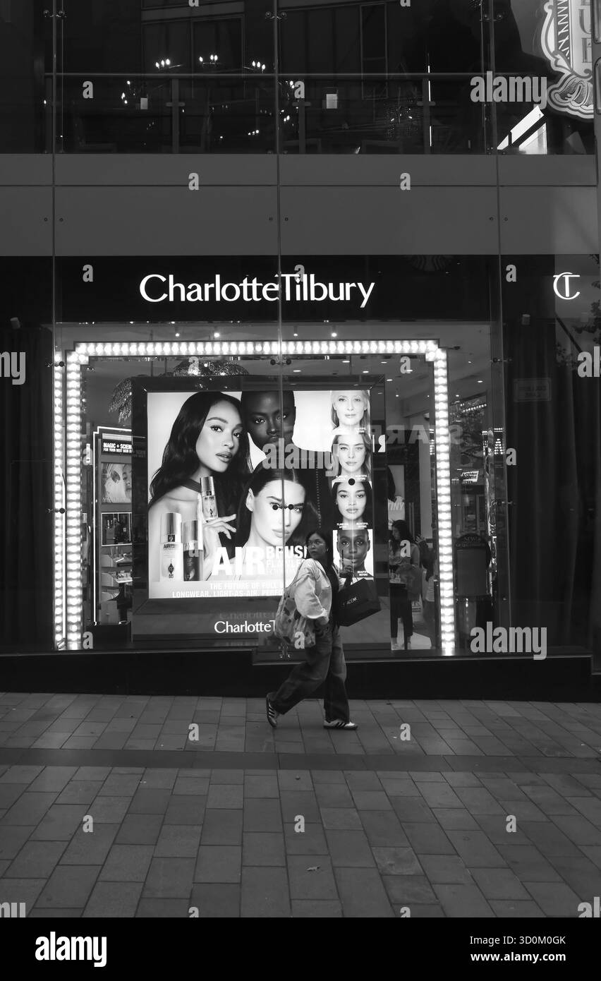 Schwarz-weiß-Foto einer Frau, die am beleuchteten Charlotte Tilbury Store in Liverpool One vorbeigeht, mit großen Schönheitsanzeigen im Fenster. Stockfoto