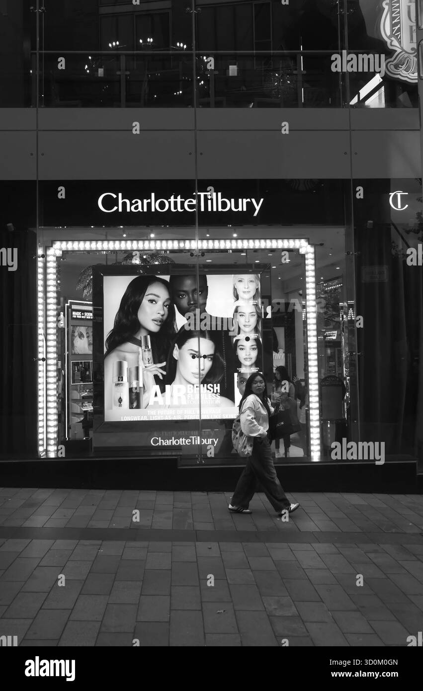 Schwarz-weiß-Foto einer Frau, die am beleuchteten Charlotte Tilbury Store in Liverpool One vorbeigeht, mit großen Schönheitsanzeigen im Fenster. Stockfoto